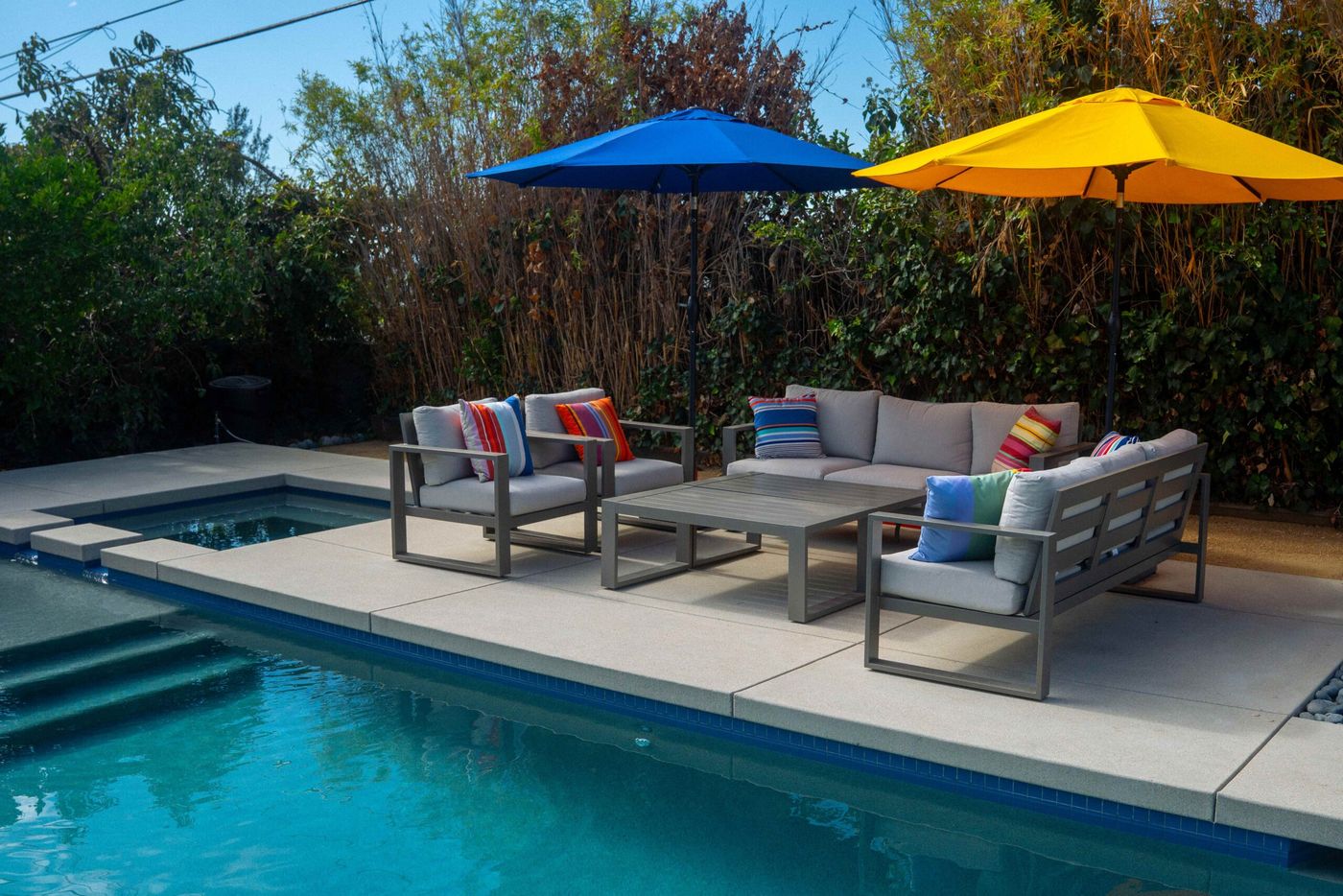Cushioned outdoor sofa and chairs beside a residential swimming pool with umbrellas and concrete decking.