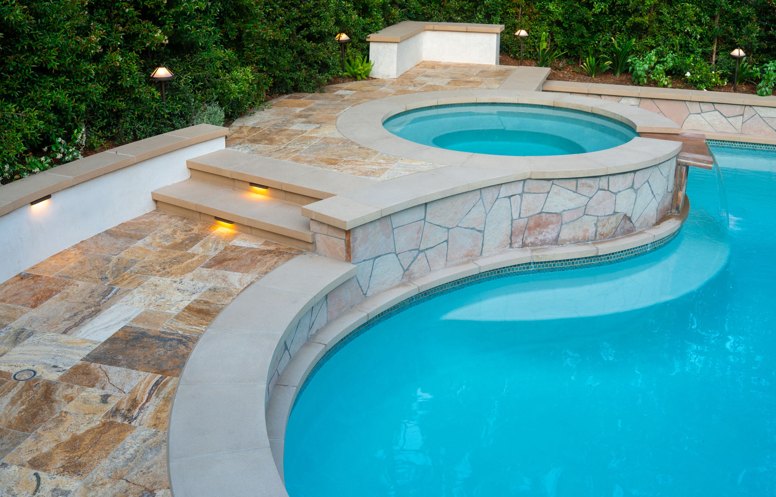 Circular raised spa with stone veneer surround, integrated steps, and adjacent swimming pool bordered by natural stone decking and landscape lighting.