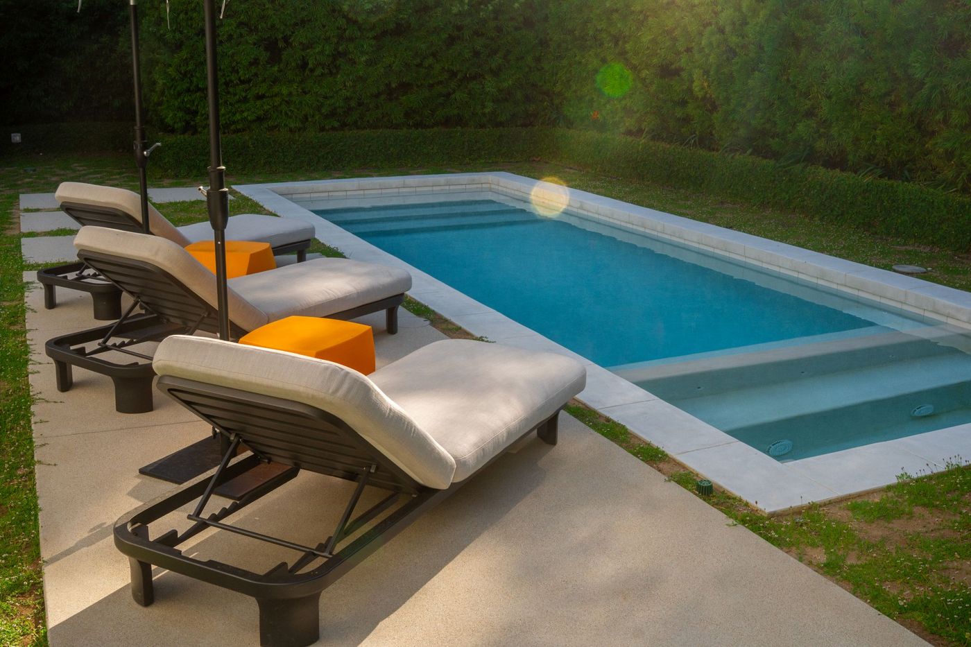 Minimalist rectangular pool bordered by concrete decking and cushioned lounge chairs in a modern backyard.