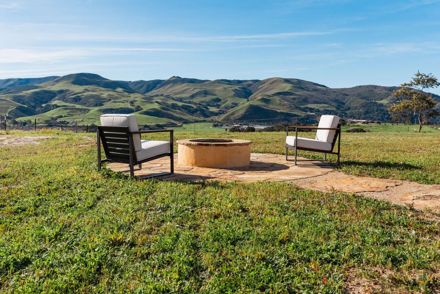 Two lounge chairs beside a stone fire pit set in an expansive open grassy field with rolling rural hills in the background