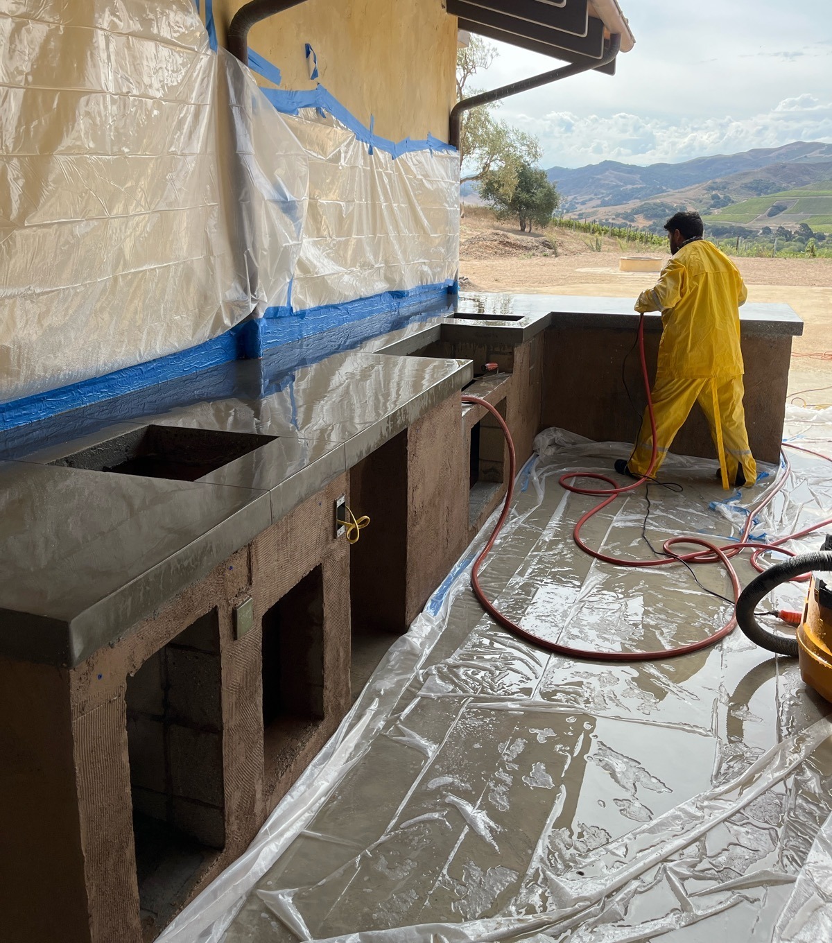 Worker sealing new concrete outdoor kitchen countertops during construction with a vineyard hillside beyond