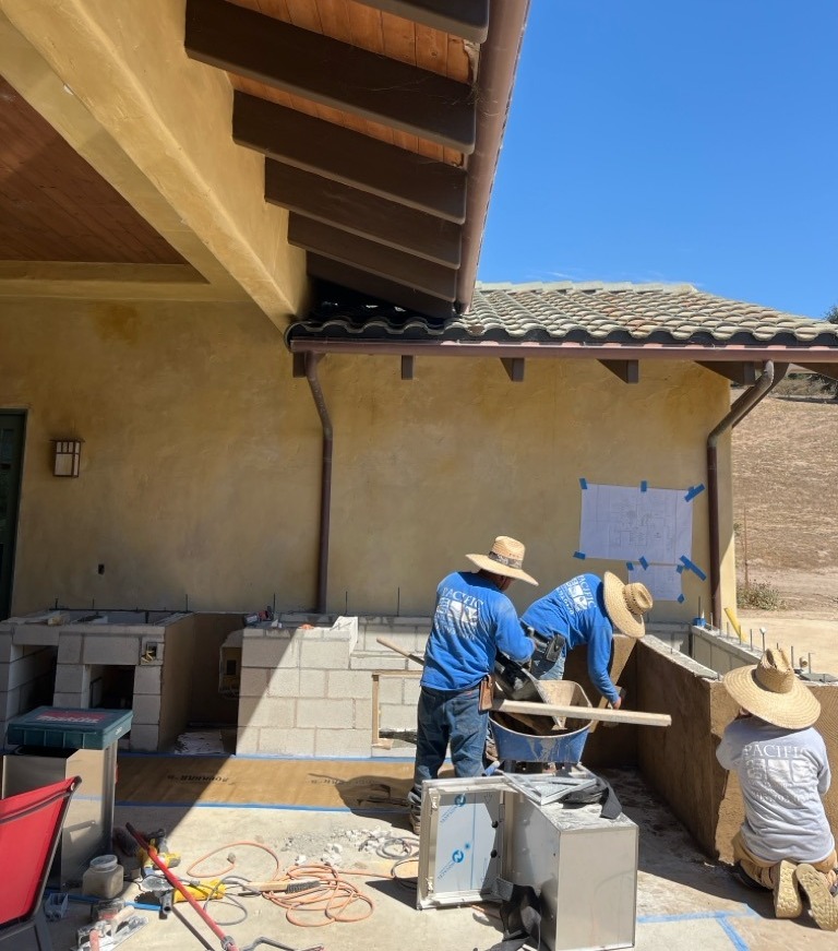 Crew building the block frame of an outdoor kitchen on a concrete patio from a taped-up plan