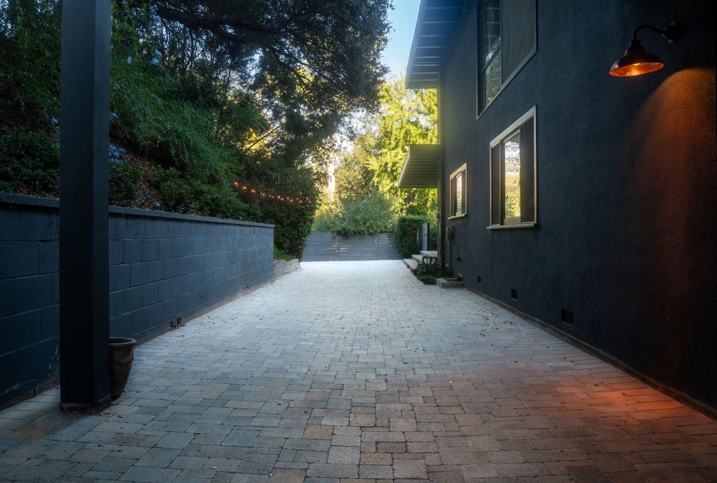 Long paver walkway along a contemporary dark-painted home with retaining wall, exterior lighting, and surrounding trees forming a clean modern landscape pathway.