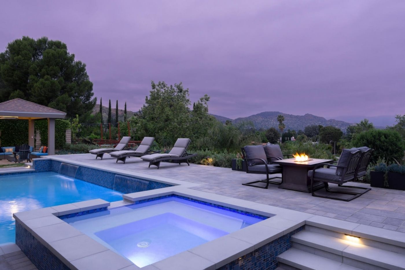 A poolside spa and fire pit table at dusk with a mountain view in the background