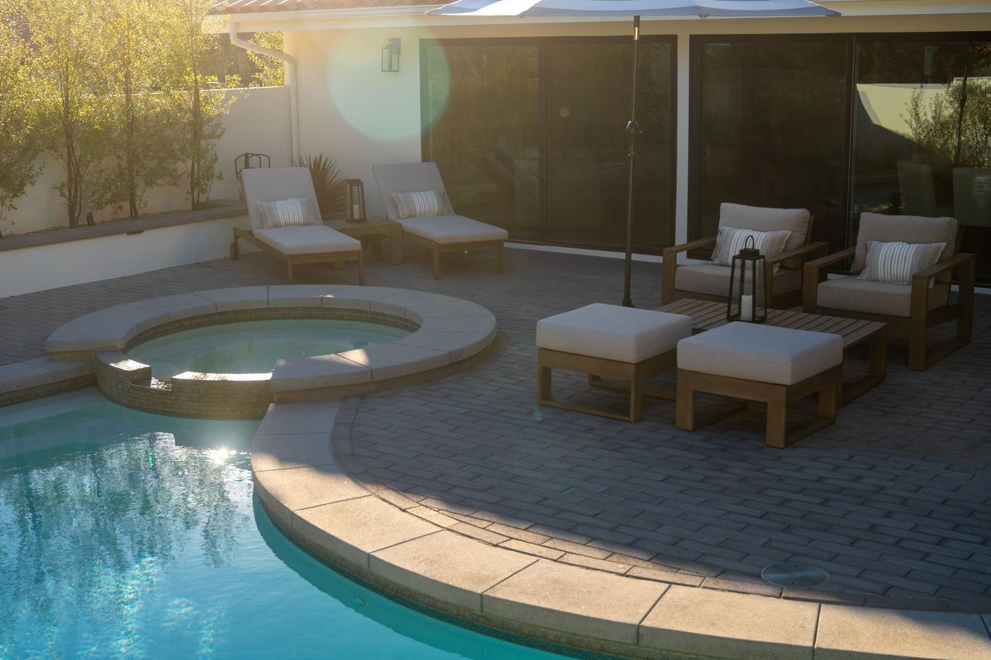 Raised spa connected to swimming pool with cushioned lounge chairs, umbrella, and paved patio in a contemporary backyard.
