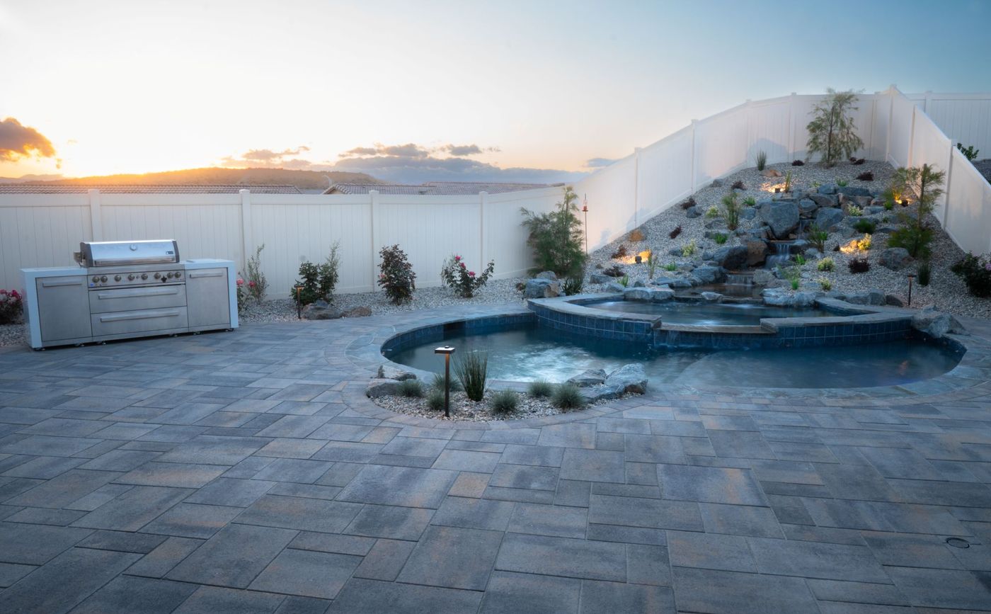 Backyard pool and raised waterfall feature beside a built-in stainless outdoor kitchen, photographed at sunset with warm sky tones