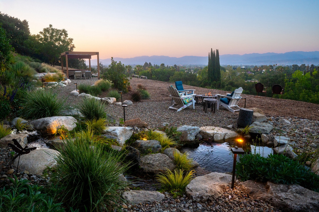 Hillside entertaining patio at dusk with fire pit seating, a pergola, and a boulder-edged pond
