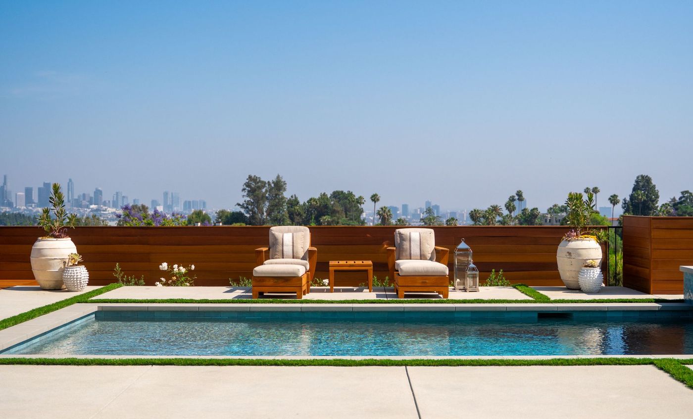 Balanced arrangement of outdoor lounge chairs facing a modern swimming pool with panoramic city skyline beyond a wood privacy fence.
