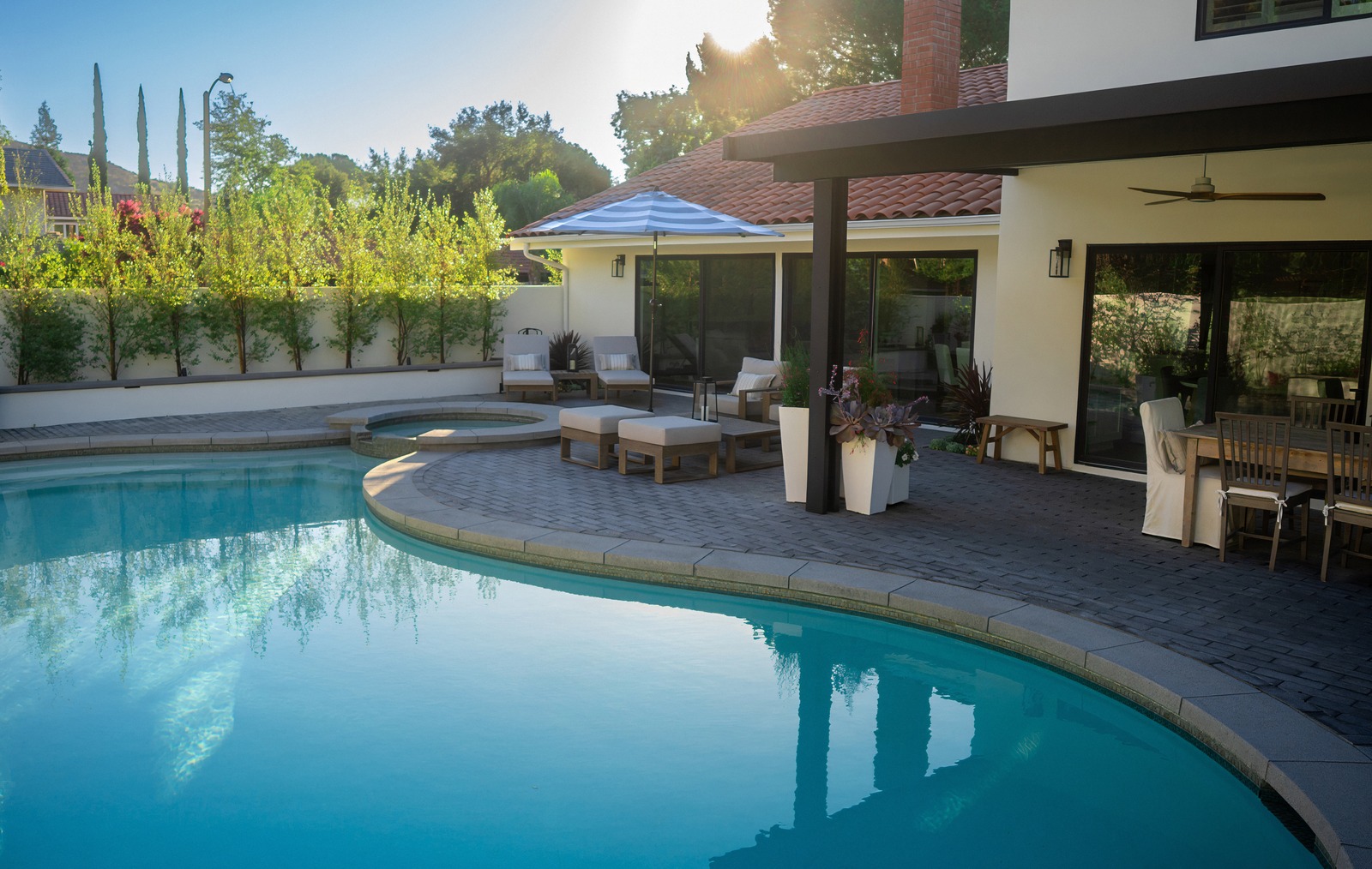 Freeform pool and raised spa edged in pavers beside a covered patio with a dining set and ceiling fan