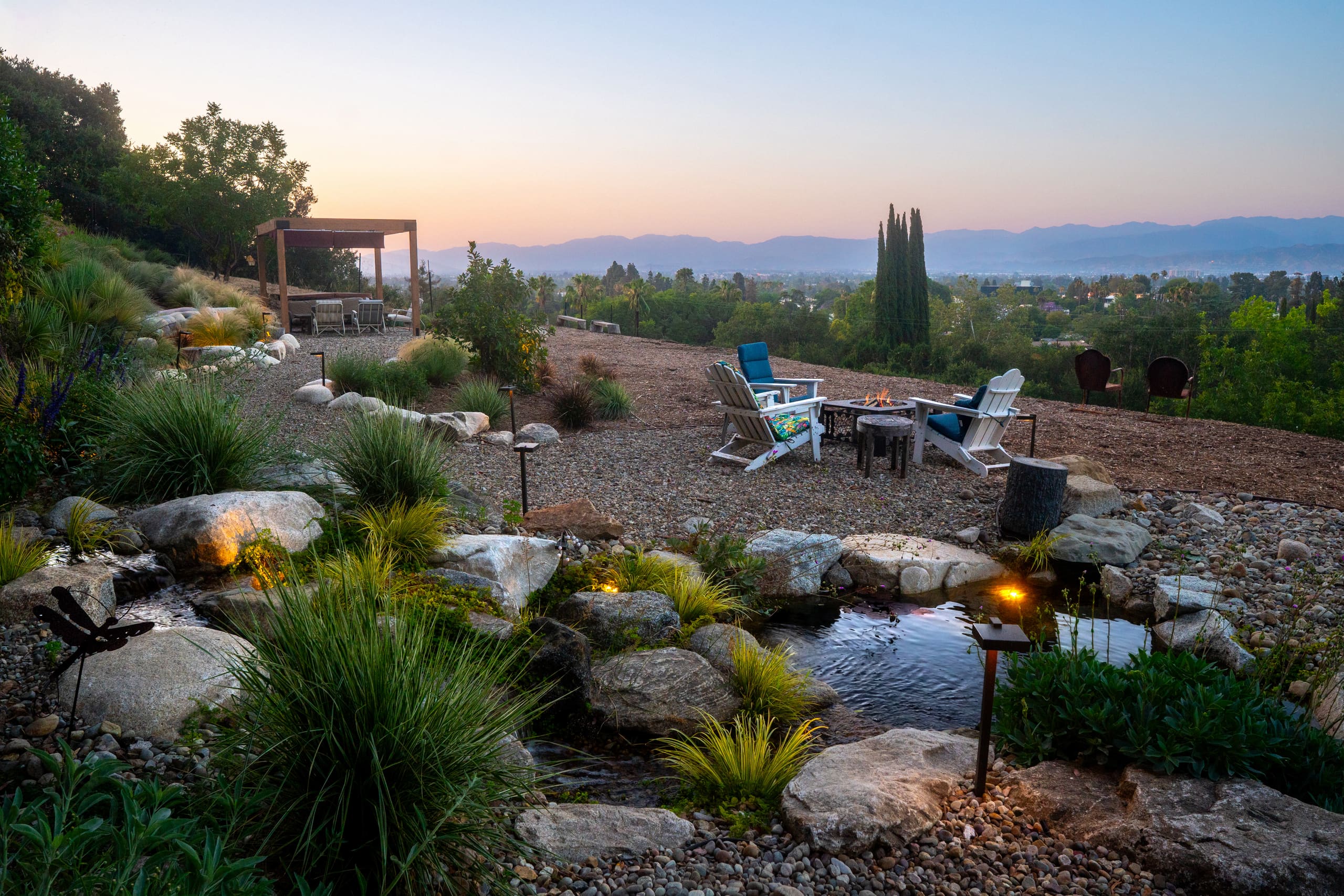 A hillside garden at dusk designed by Julian Montford, with a lit fire pit and chairs, a timber pergola and a boulder-lined creek above a valley view
