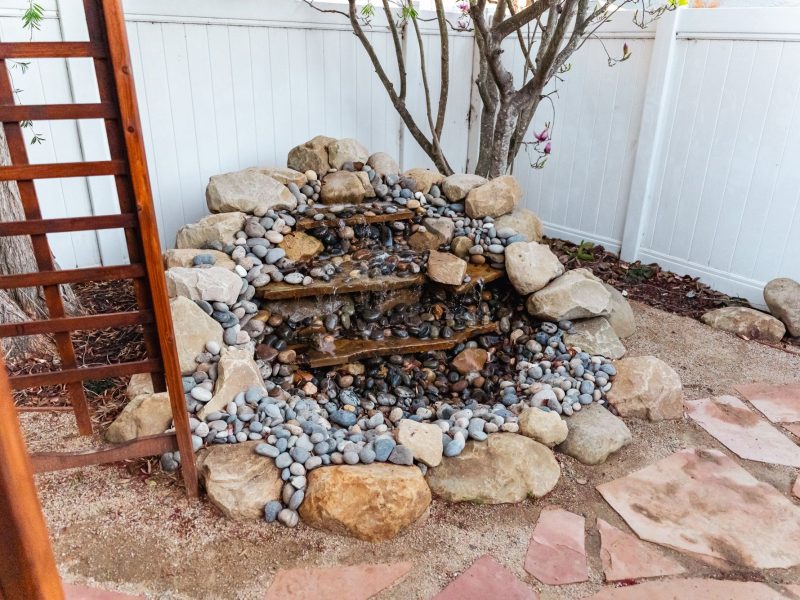 Close-up of tiered natural stone waterfall surrounded by river rock in a backyard patio setting.