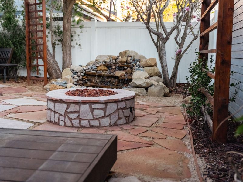 Custom backyard flagstone patio featuring a circular stone fire pit and natural rock waterfall water feature.