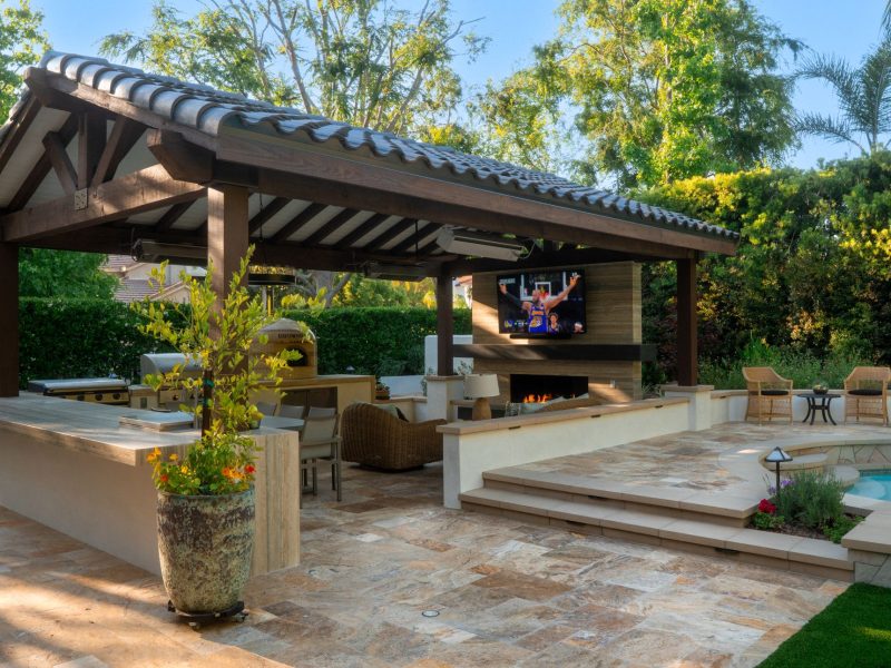 Wide view of covered outdoor kitchen pavilion, fireplace lounge, stone patio, and adjacent swimming pool creating a resort-style backyard retreat.