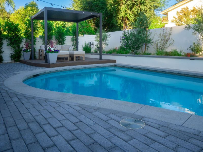 Residential swimming pool with curved edge, pergola lounge seating, landscaping, and paver patio in a private backyard.