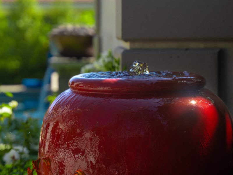 Red ceramic bubbling fountain surrounded by garden plants in a landscaped backyard near a swimming pool.