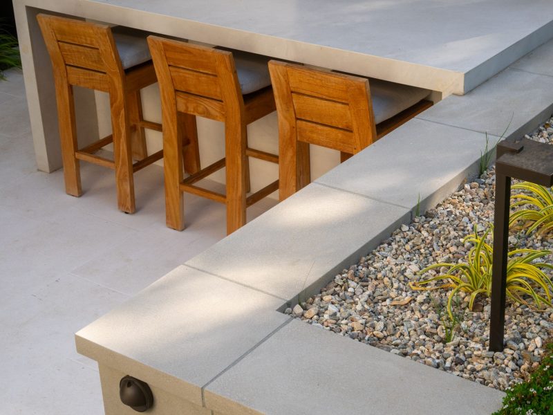 Wood bar stools tucked beneath a sleek concrete outdoor kitchen counter with decorative gravel landscape.