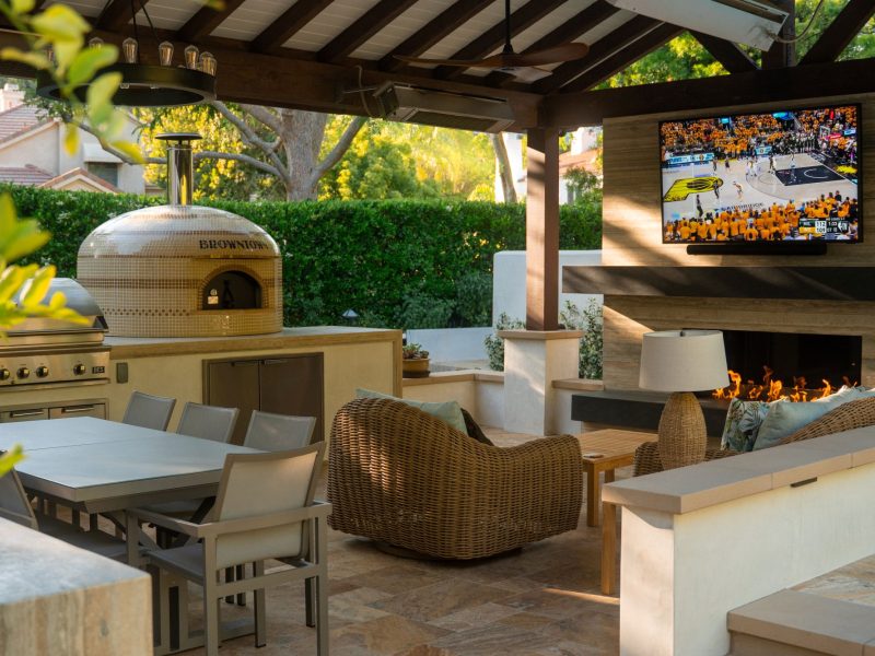 Covered outdoor living space featuring built-in grill, pizza oven, dining area, fireplace with mounted TV, and comfortable lounge seating in a landscaped backyard.
