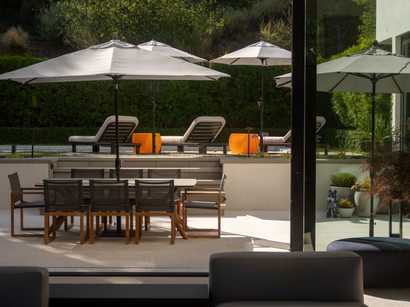 Shaded outdoor dining patio with table and chairs overlooking a landscaped backyard and pool lounge area.