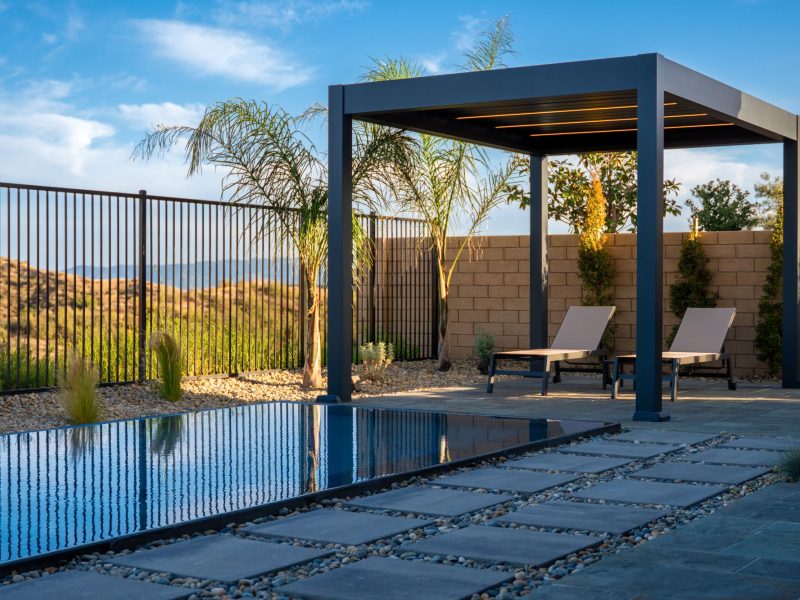 Wide view from beneath pergola showing outdoor dining, linear pool, stepping stone path, and expansive sky over contemporary backyard design.