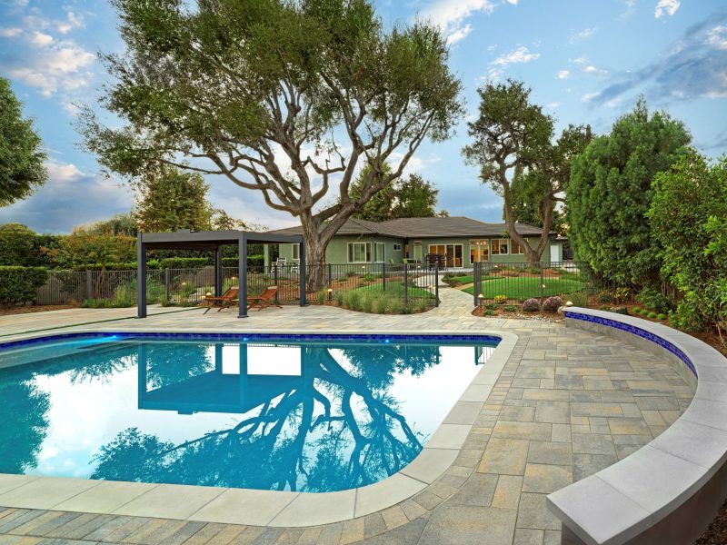 Wide view of a modern backyard with curved pool coping, paver deck, mature trees, lawn areas, and pergola lounge creating a resort-style outdoor environment.