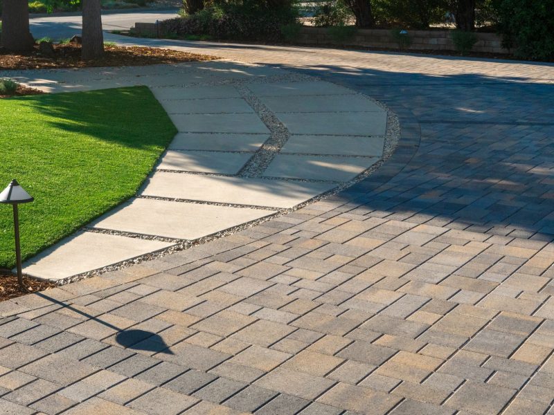 Smooth curved concrete walkway connecting lawn area to patterned paver driveway with integrated landscape lighting in a modern residential landscape.