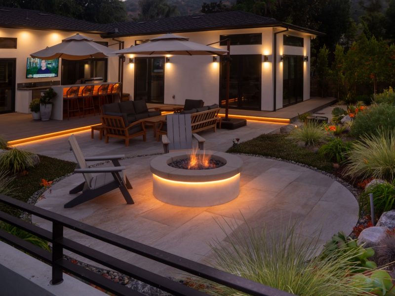 Contemporary backyard at dusk with illuminated fire pit, patio lighting, outdoor seating, and modern home architecture.