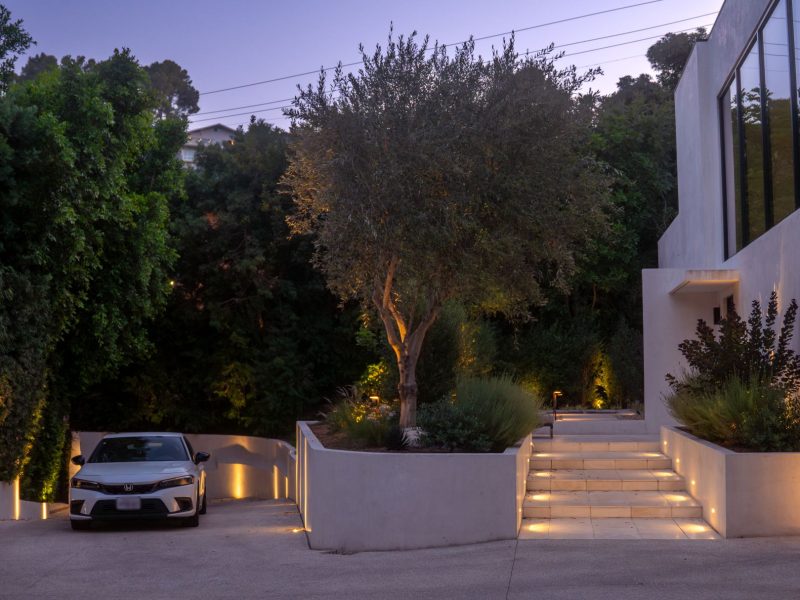 Contemporary home entry with illuminated steps, driveway lighting, and mature trees at dusk.