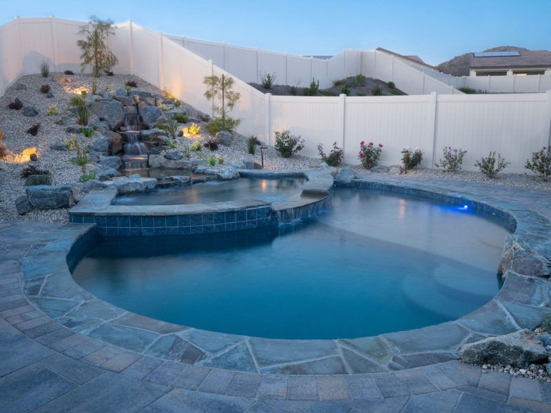 Freeform swimming pool featuring natural rock waterfall, raised spa edge, and soft landscape lighting during evening twilight.