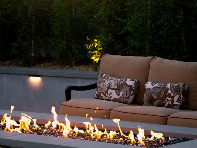 Modern linear fire pit with glass media in front of cushioned outdoor seating and landscape lighting.