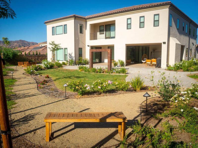 Wide view of landscaped backyard courtyard featuring curved decomposed granite pathways, lawn, flowering plants, and contemporary two-story home architecture.