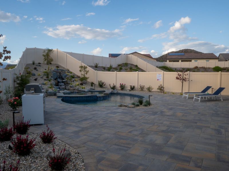 Custom backyard swimming pool with rock waterfall, large paver patio, and modern outdoor lounge area in a landscaped residential yard.