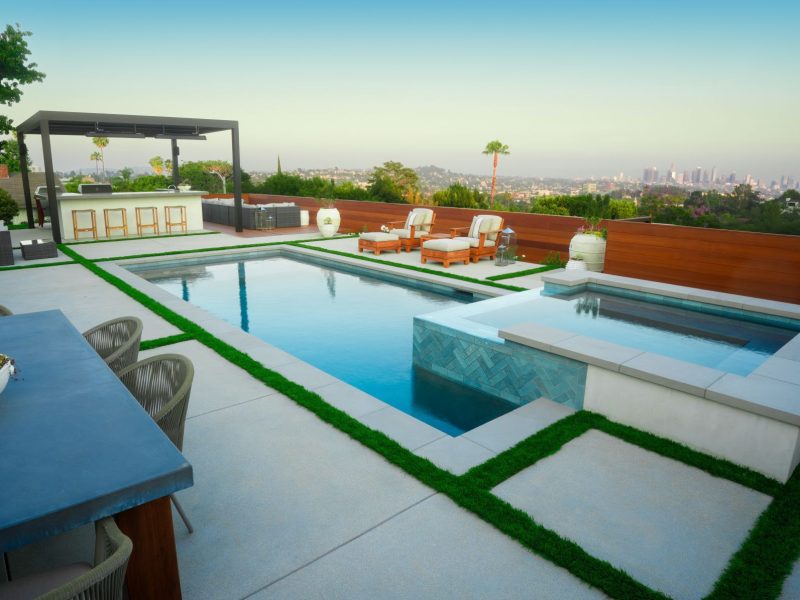 High-end backyard design featuring geometric swimming pool, raised spa, artificial turf joints, pergola outdoor kitchen, and city skyline views.