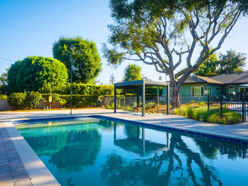 Wide view of contemporary backyard pool, pergola lounge, paver deck, fencing, and surrounding greenery creating a cohesive California outdoor living space.