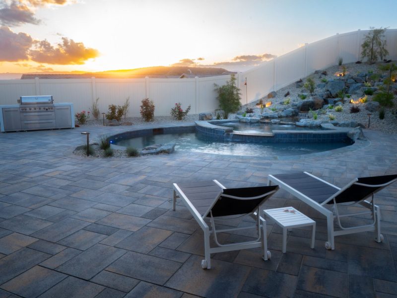 Luxury residential poolscape at sunset featuring freeform pool, illuminated rock waterfall, wide paver patio, and contemporary lounge chairs.