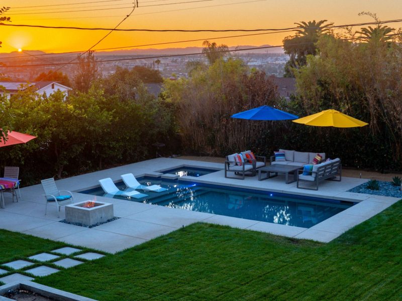 Wide view of contemporary backyard with swimming pool, spa, fire feature, lounge seating, and sunset skyline.