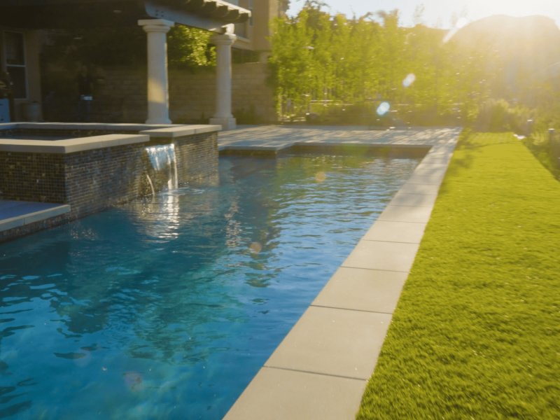 Long rectangular lap pool beside green lawn and raised spa with waterfall in a sunlit luxury backyard.
