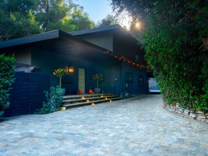 Wide residential paver driveway leading to a modern home with integrated step lighting, string lights, and lush surrounding greenery in a California outdoor living design.