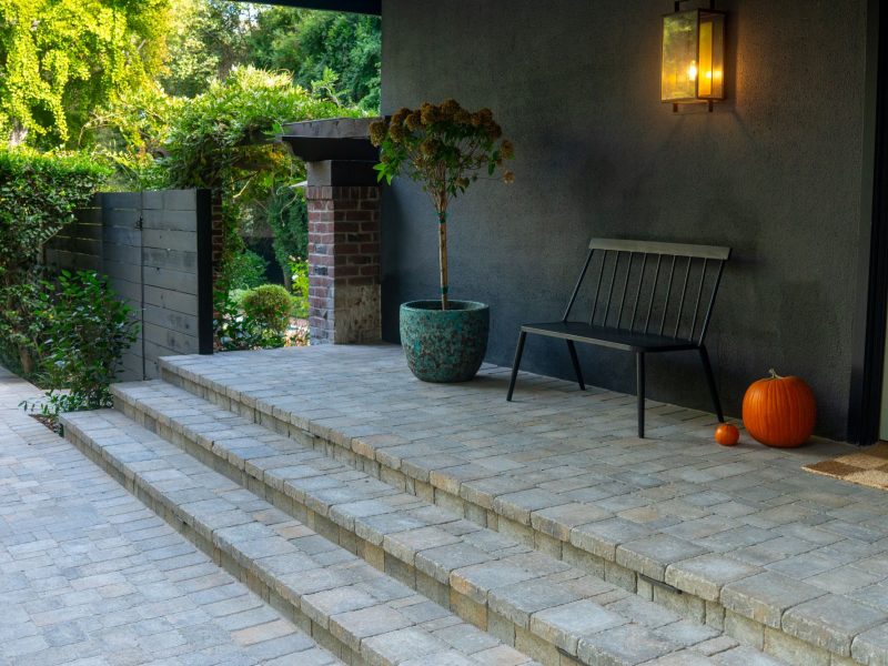 Contemporary front porch with multi-level paver steps, dark exterior walls, decorative planters, and warm entry lighting creating an inviting modern landscape design.