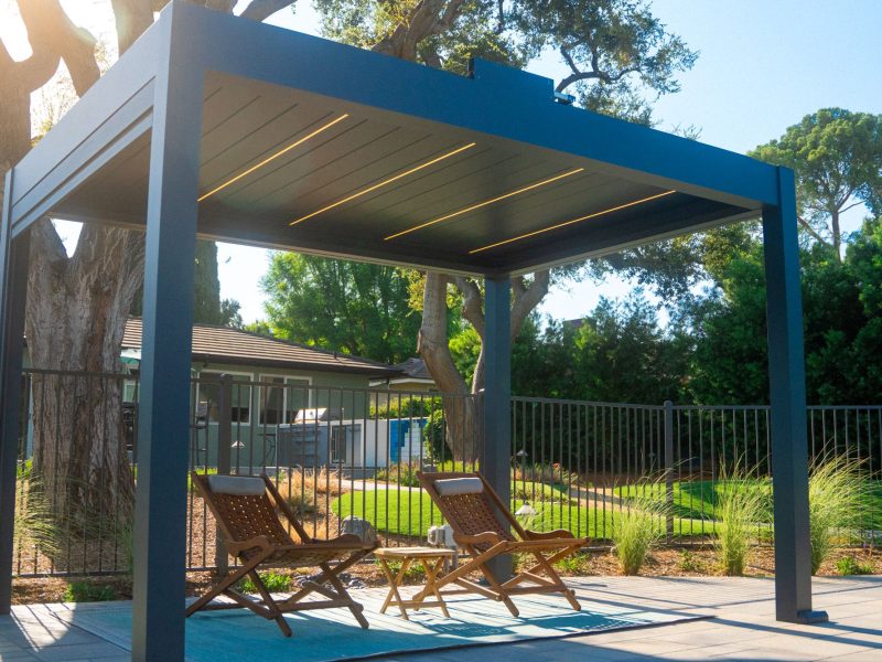 Contemporary backyard pergola with integrated linear lighting, lounge chairs, and surrounding greenery creating a shaded outdoor living retreat in a California landscape design.