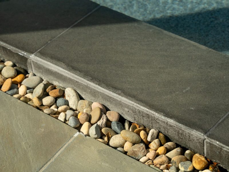 Close view of smooth dark pool coping beside natural river rock drainage channel highlighting clean minimalist pool design detail.