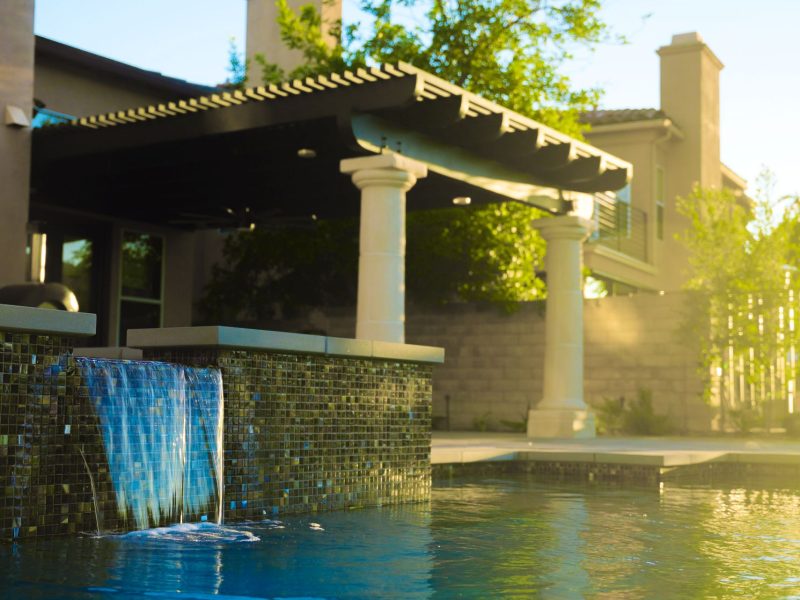 Contemporary lap pool featuring raised spa, waterfall, covered pergola patio, and landscaped yard.
