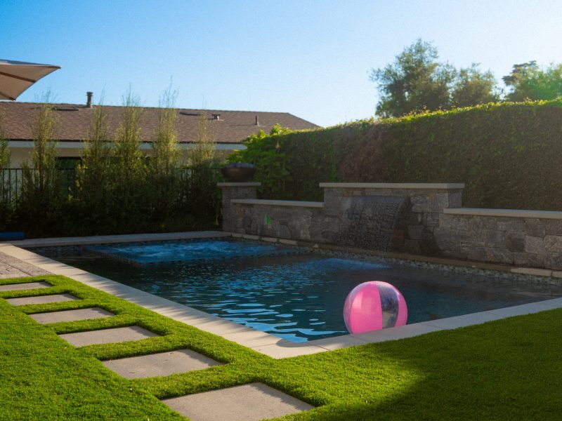 Rectangular backyard swimming pool with green lawn, concrete stepping stones, and stone wall water feature.