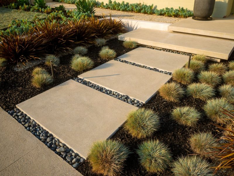 Close-up of contemporary concrete stepping stone walkway with decorative river rock joints, drought-tolerant ornamental grasses, and clean modern landscape design.