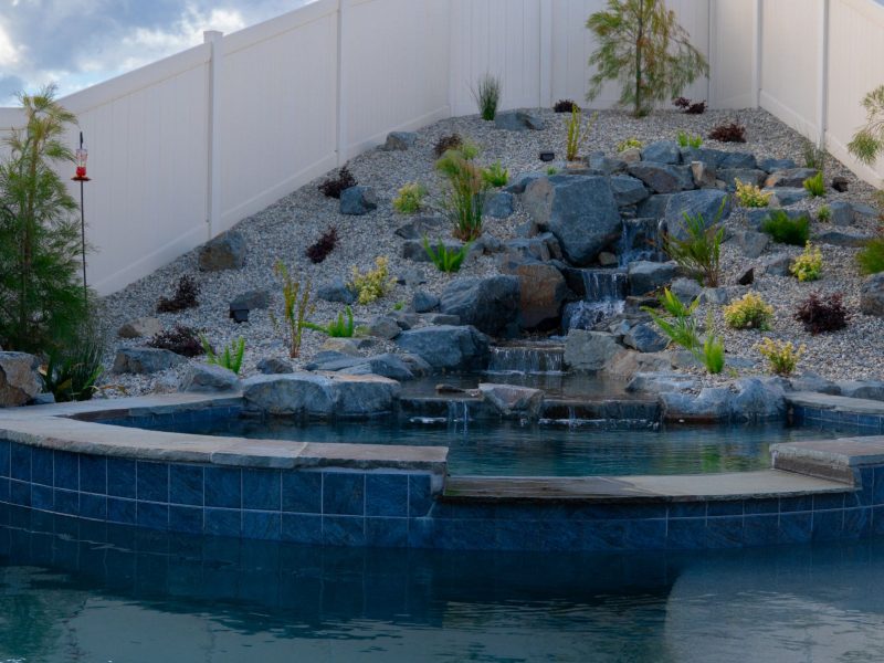 Close-up of raised spa edge with cascading natural stone waterfall flowing into a custom swimming pool surrounded by gravel landscaping.