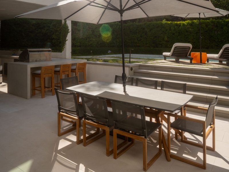 Modern patio dining space connected by steps to elevated poolside lounge chairs and landscaped greenery.