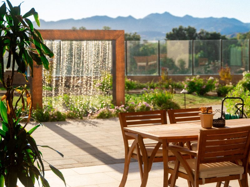 Patio dining table and chairs positioned beside a decorative water wall feature and landscaped courtyard garden with mountain backdrop.