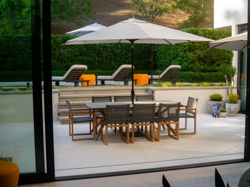 View through sliding glass doors showing outdoor dining patio, steps, and pool lounge chairs in modern backyard.