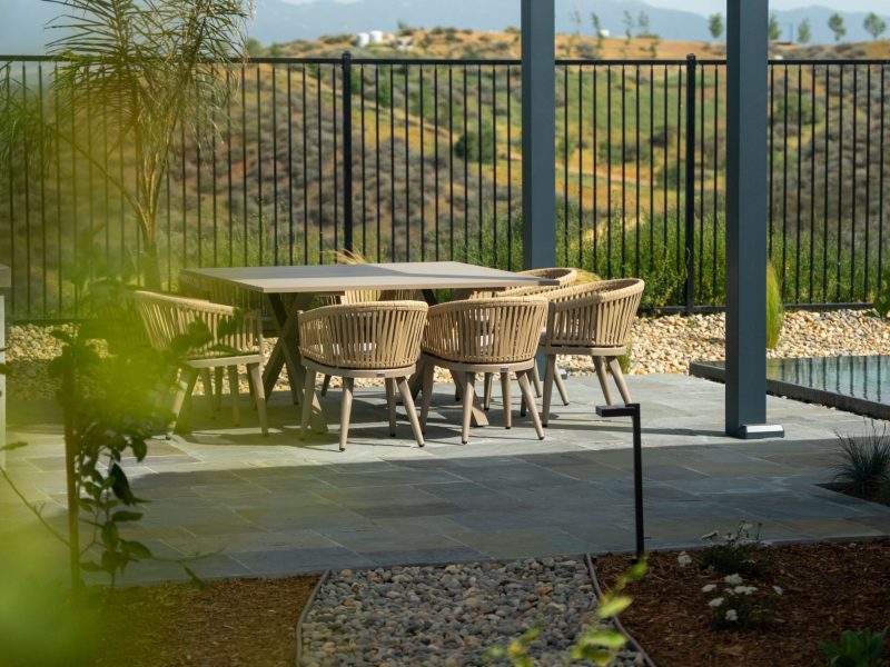 Outdoor dining table and woven chairs beneath sleek modern pergola next to long pool with hillside landscape backdrop.