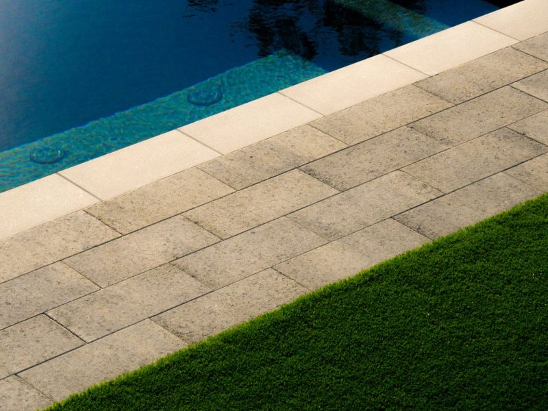 Close-up of swimming pool coping, stone pavers, and artificial turf in a modern landscaped backyard.