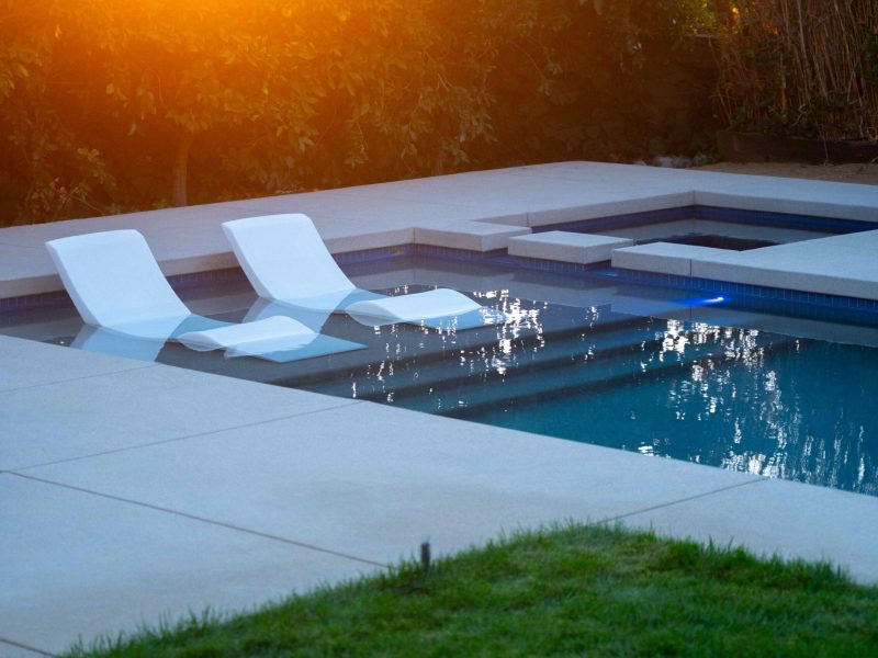 Swimming pool tanning shelf with white lounge chairs illuminated by warm sunset light in a modern backyard.